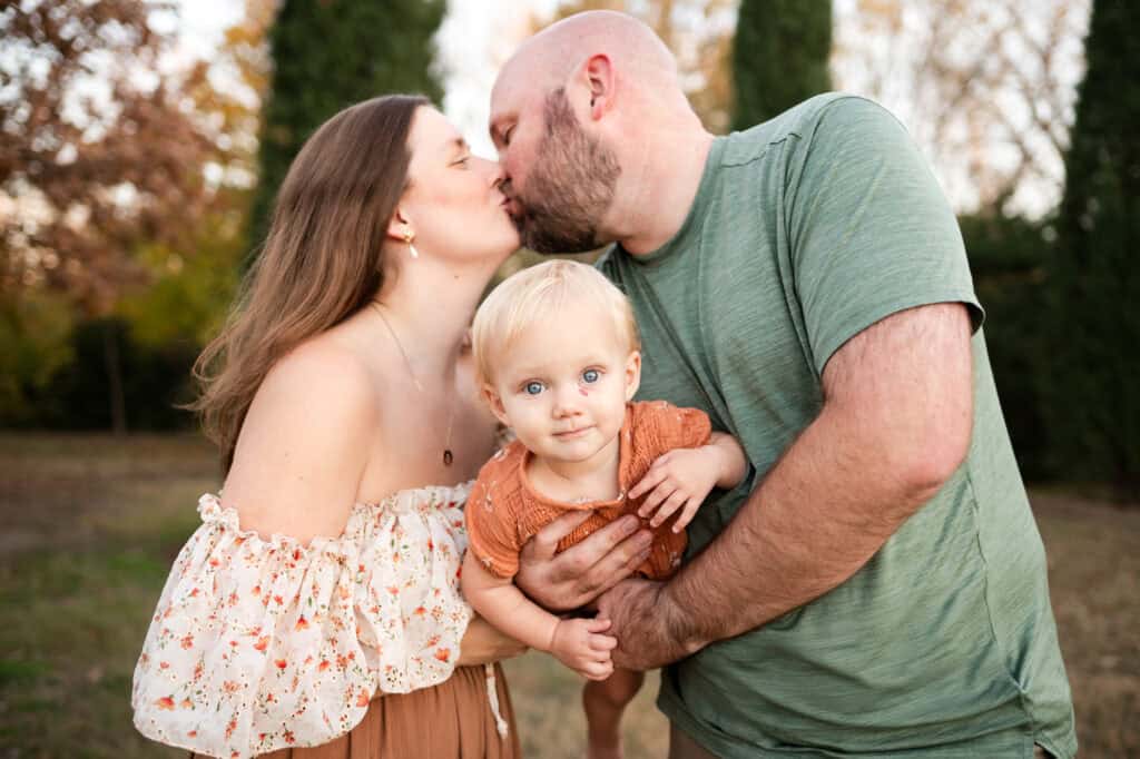 parents kiss holding baby girl in sedgwick county park with wichita family photographer sj nuzum photography